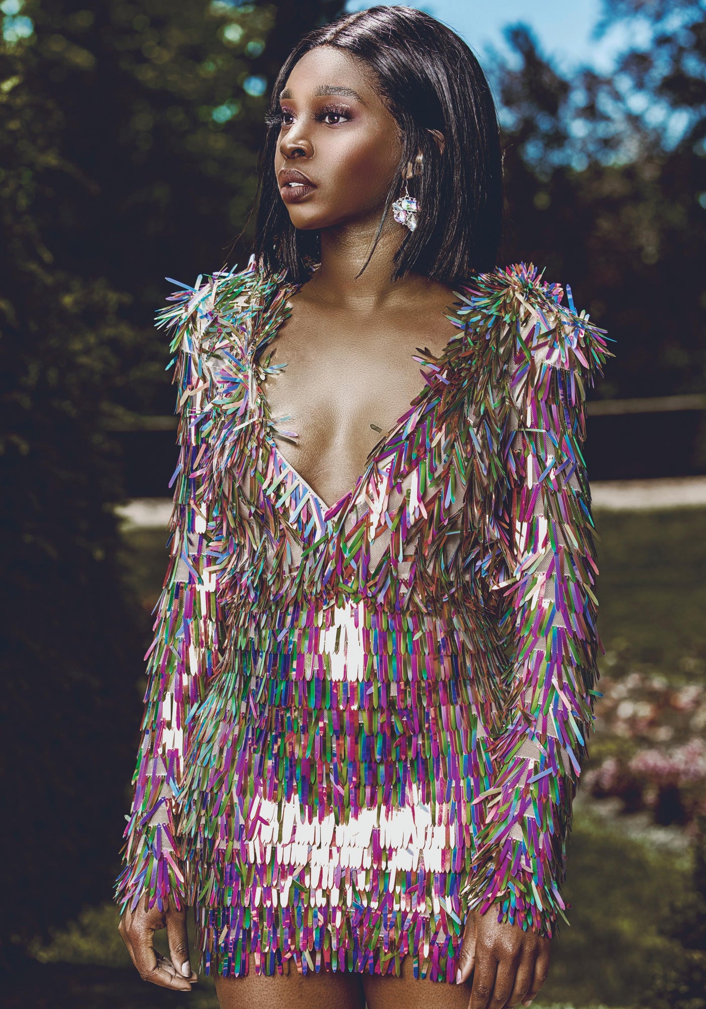 Model in a deep V-neck iridescent fringe mini dress, shimmering in the sunlight against a lush backdrop.