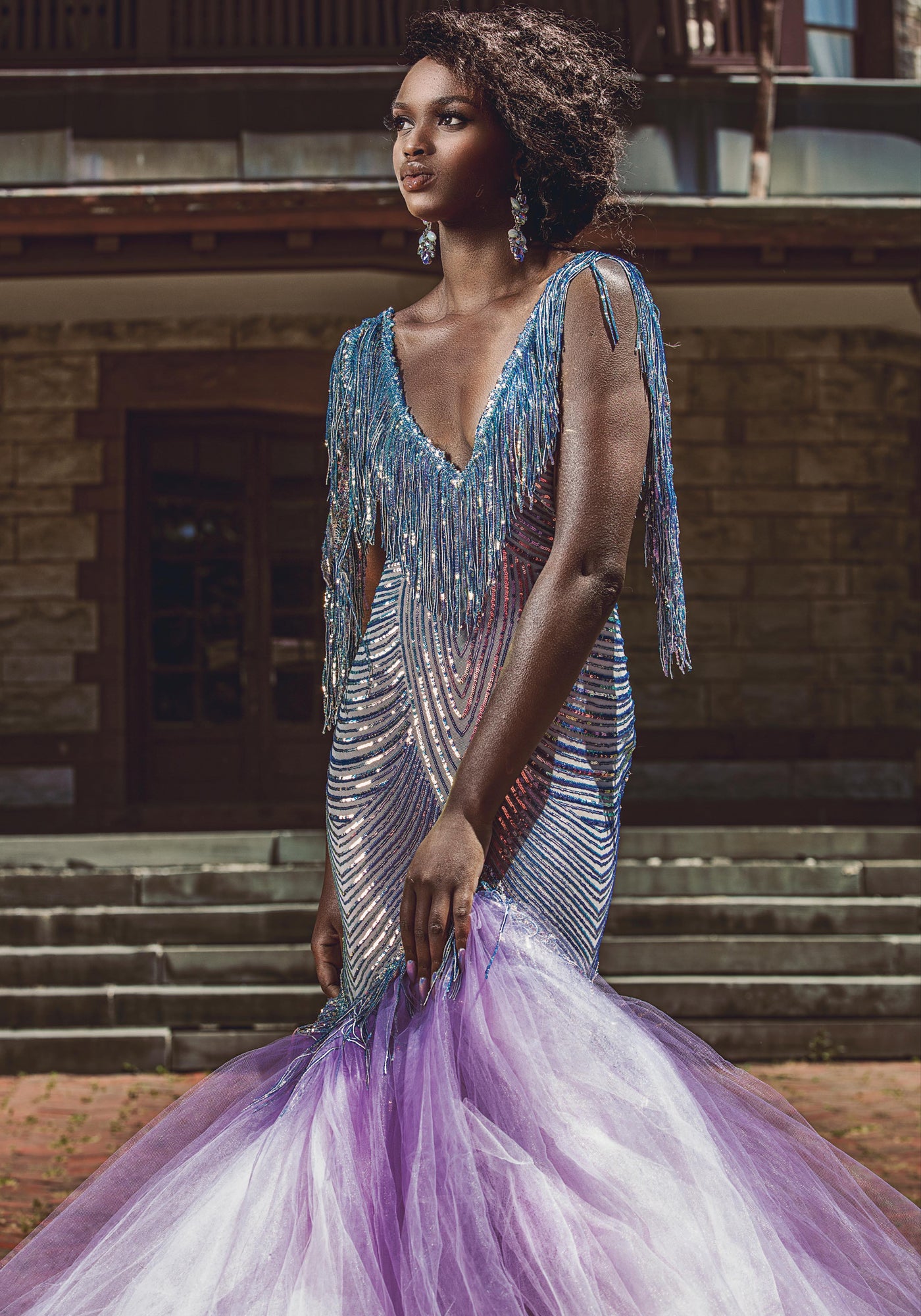 Model in a shimmering blue and lavender mermaid gown with fringe details, standing elegantly on stone steps